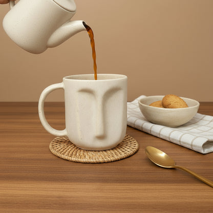 Beige face-shaped mug on a gray surface with a black mug and decorative elements in the background.