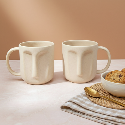 Two face-shaped mugs on a table with a bowl of cookies and a spoon.