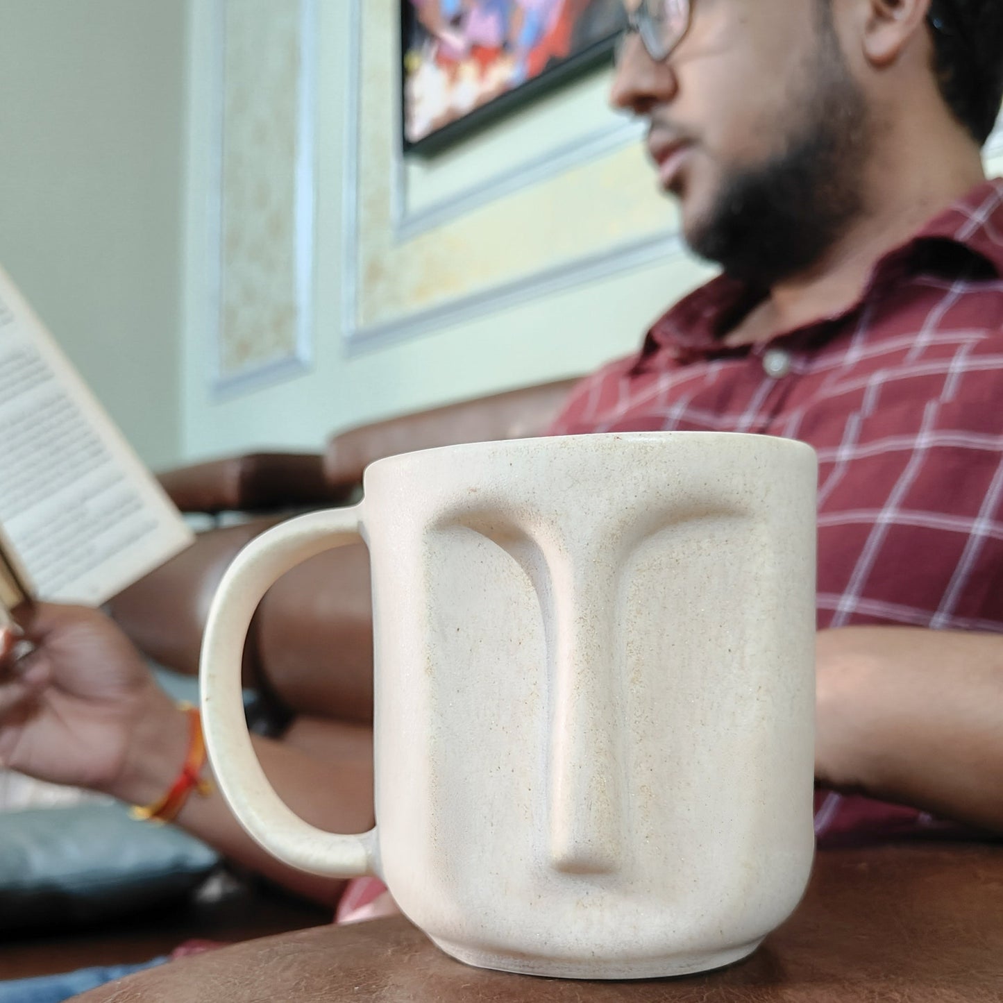 Person sitting on a couch with a ceramic mug featuring a face design, holding a newspaper.