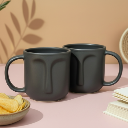 Two dark gray mugs on a wooden surface with a bowl of chips and a book in the background.