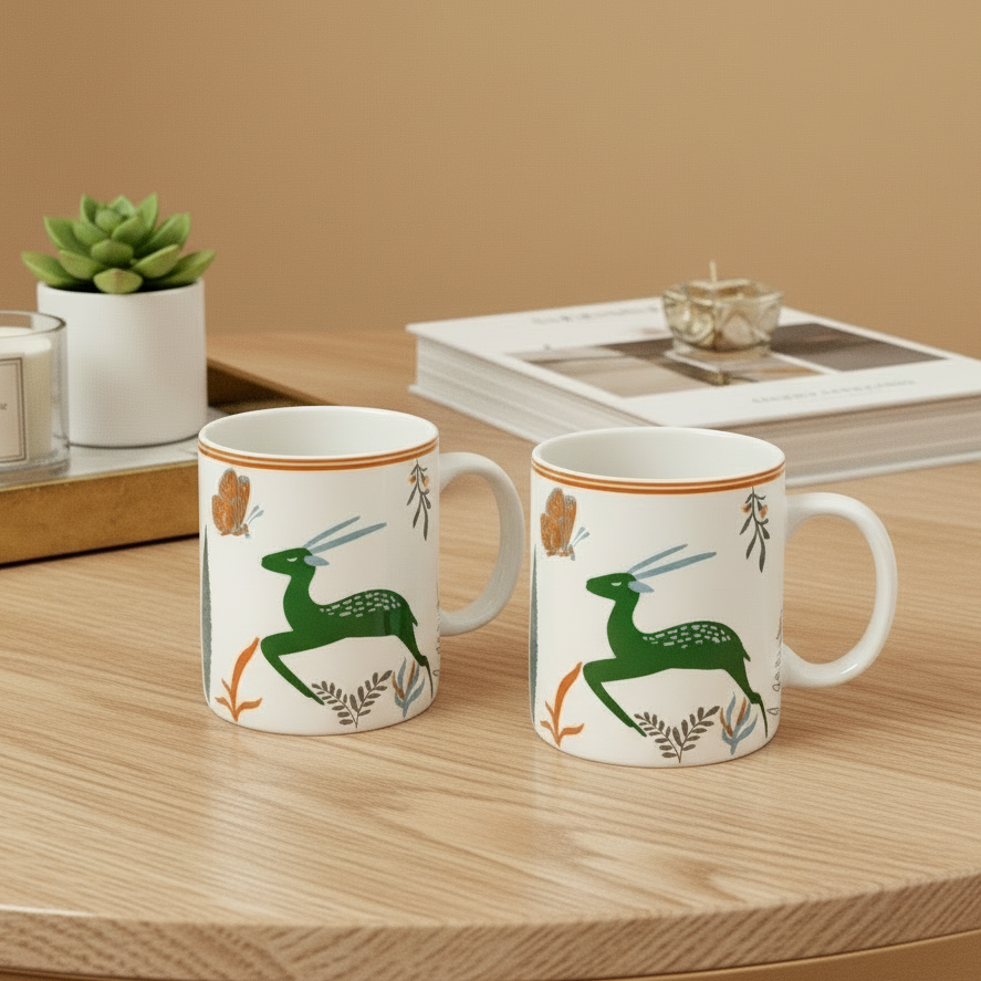 Two mugs with deer design on a marble table with a candle and plant in the background.