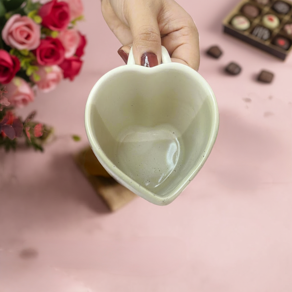 Heart-shaped ceramic cup held by a hand with pink roses and chocolates in the background
