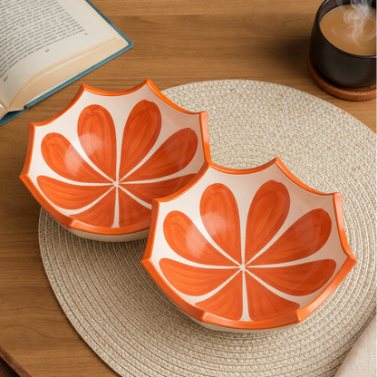 An orange and white ceramic bowl with a petal-like pattern, styled on a woven placemat on a wooden table with an open book and a cup of coffee nearby.