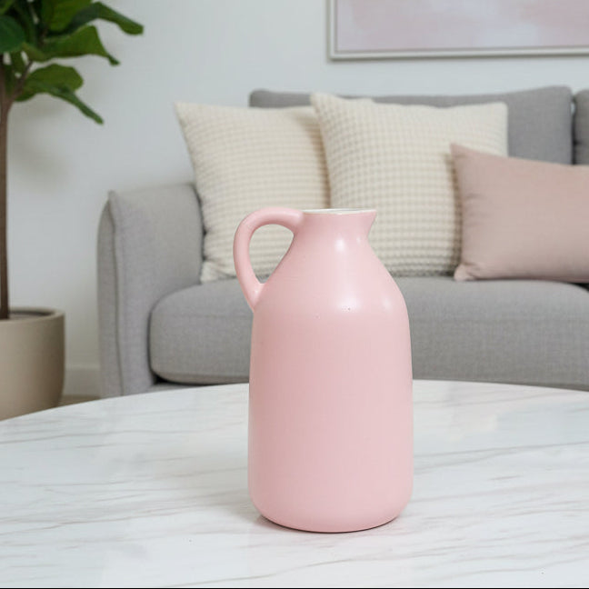 A light pink ceramic pitcher with a single handle, placed on a brown faux-leather surface with a brown leather sofa and teal pillows in the background.