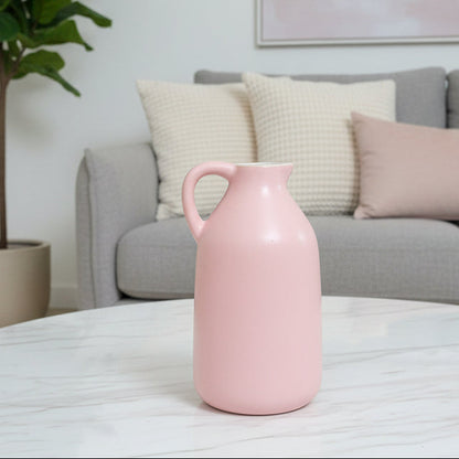 A light pink ceramic pitcher with a single handle, placed on a brown faux-leather surface with a brown leather sofa and teal pillows in the background.