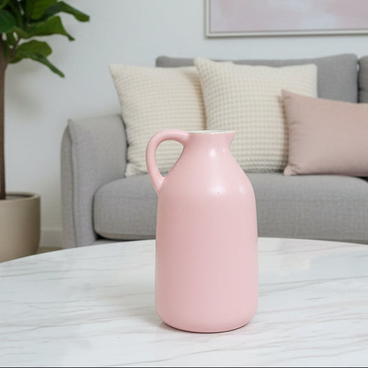 A light pink ceramic pitcher with a single handle, placed on a brown faux-leather surface with a brown leather sofa and teal pillows in the background.
